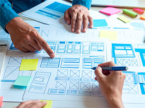 Hände arbeiten an Wireframes und Skizzen für Webseitenlayouts auf einem Tisch mit Papier, Stiften und Haftnotizen